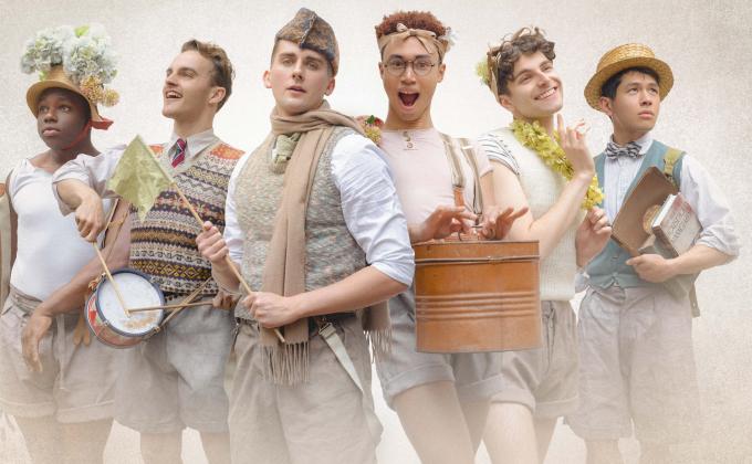 Six men stand together dressed in beige shorts and shirts, they have scarves, drums and flowered hats