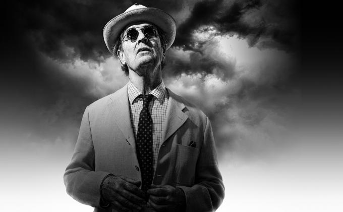 A man in a suit, hat and sunglasses stands against a cloudy sky