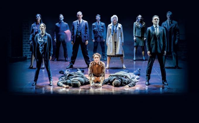 A group stand in the middle of a stage, two men lie on the ground, one each side of a woman who looks sorrowful