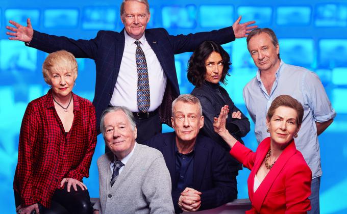 A group of people stand together around a newsdesk, one man has his arms outstretched and a woman holds her palm up against a man's face