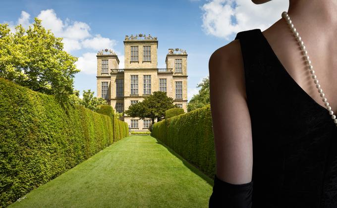 A woman in a black dress and pearls stands in front of a mansion