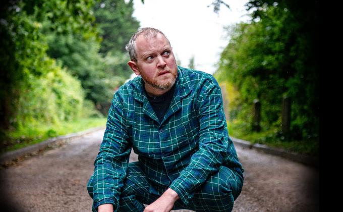 Miles Jupp crouching on a path in a green plaid shirt