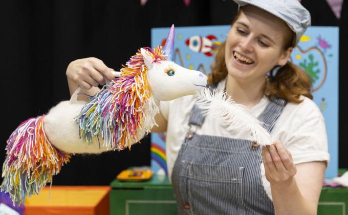 A woman wearing dungarees and a hat holds a unicorn and a big feather