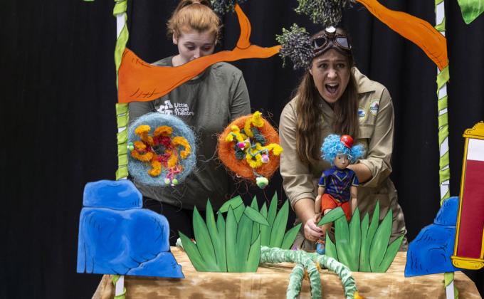 Two puppeteers in a jungle scene, one puppeteer is holding a puppet with blue hair