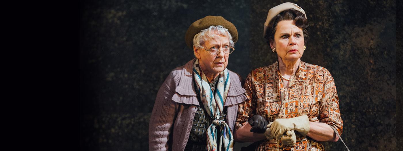 Two older woman with scarfs, hats and handbags look to the distance a little afraid.