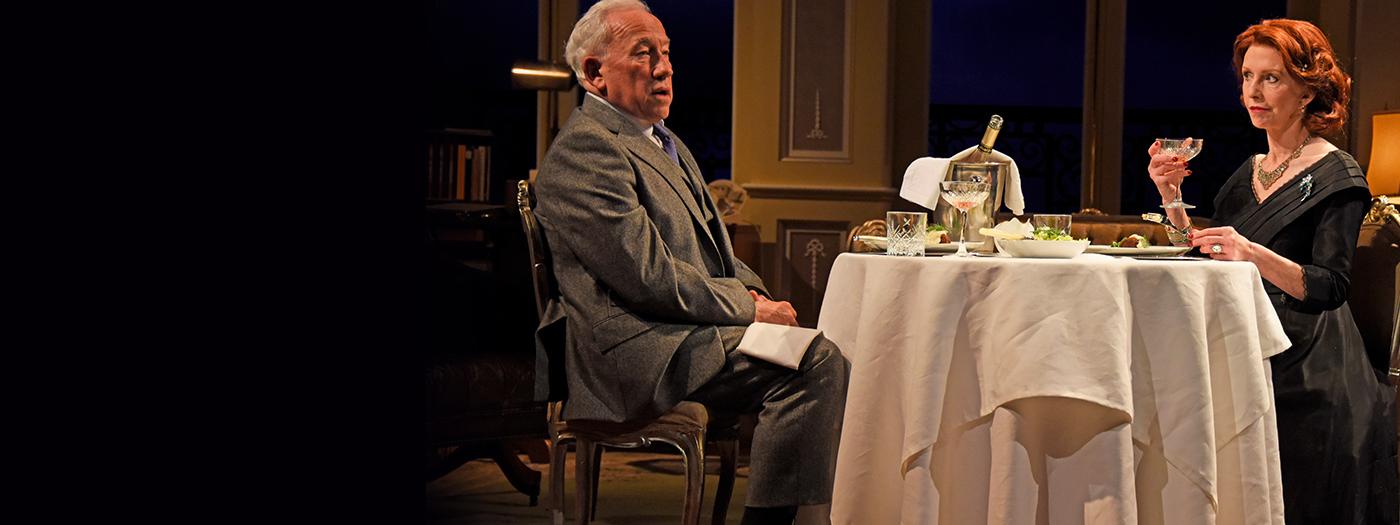 A couple sit at a dining table facing each other, one holds a drink. They both look fed up.