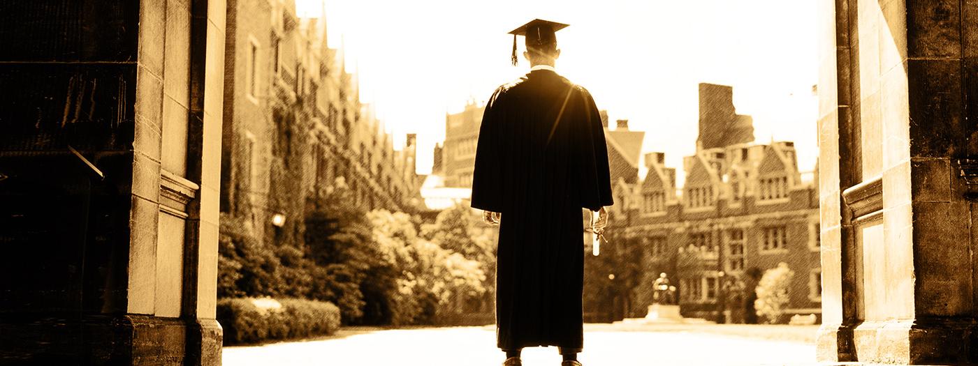 A boy in a cap and gown stands in front of a university college building