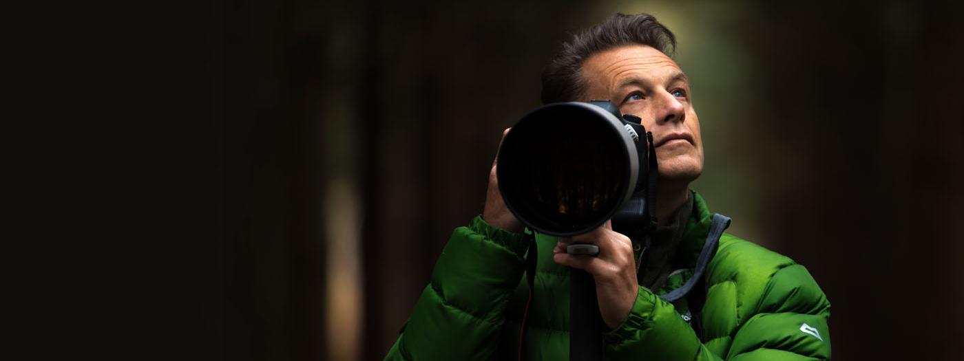 A man in a green puffer jacket holds a camera, he is standing in a forest