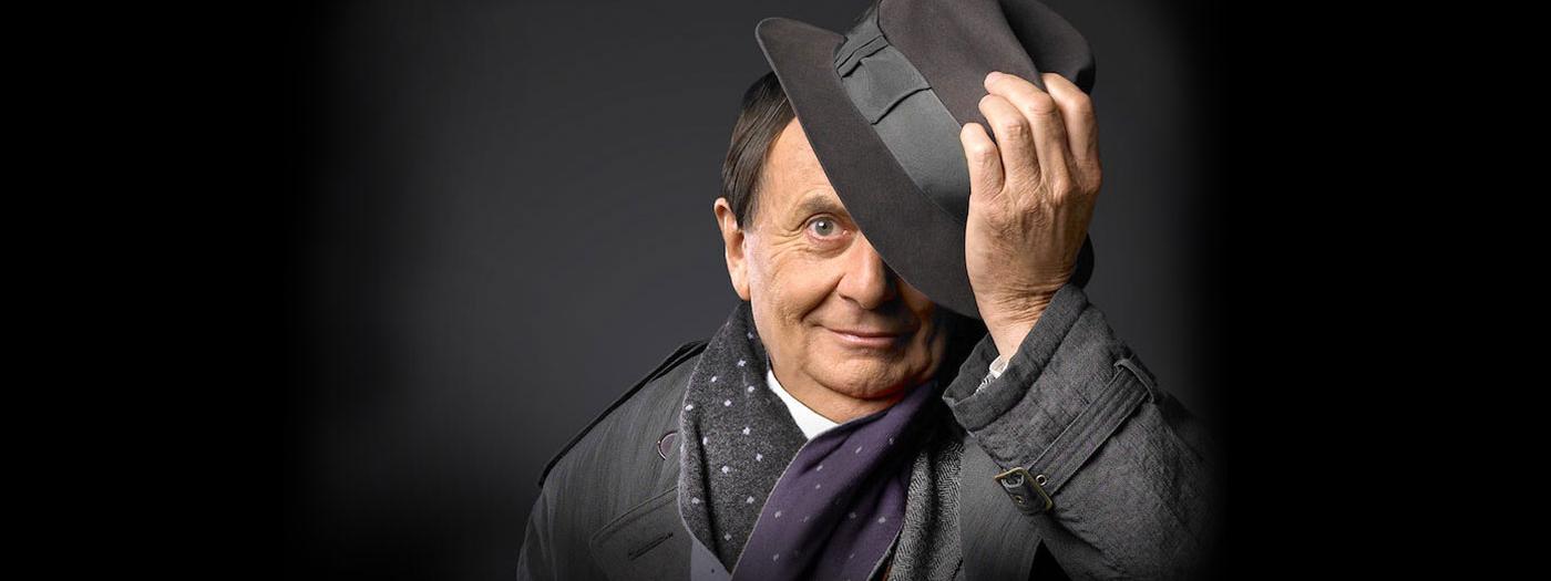 Barry Humphries wears a dark suit and covers half of his face with a hat