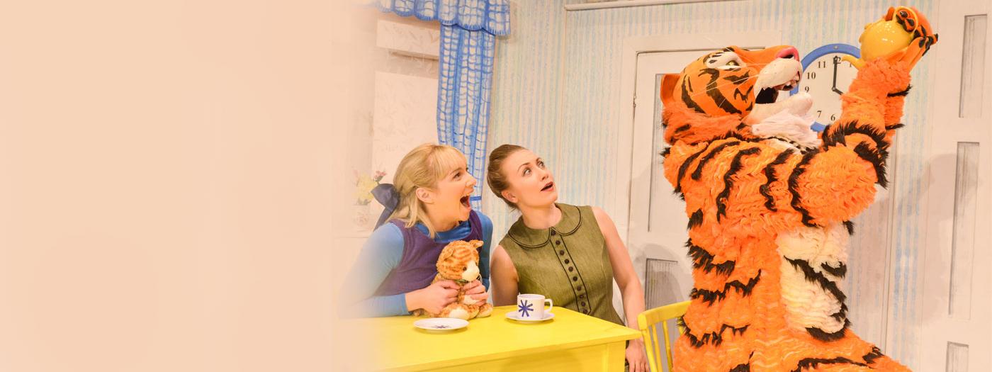 A young girl and her mother sit at a kitchen table, staring as a big Tiger drinks from a teapot
