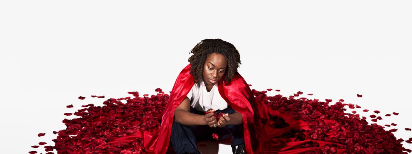 A woman sits on the floor in a red cape, which turns into petals on the floor