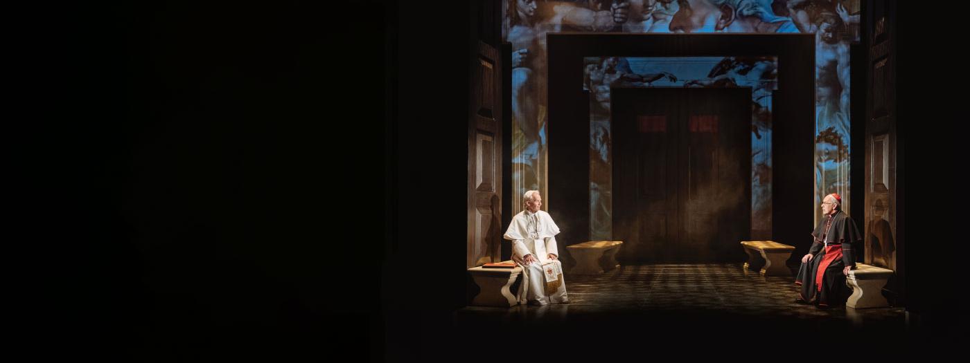 Two popes sit opposite each other on benches in a grand room