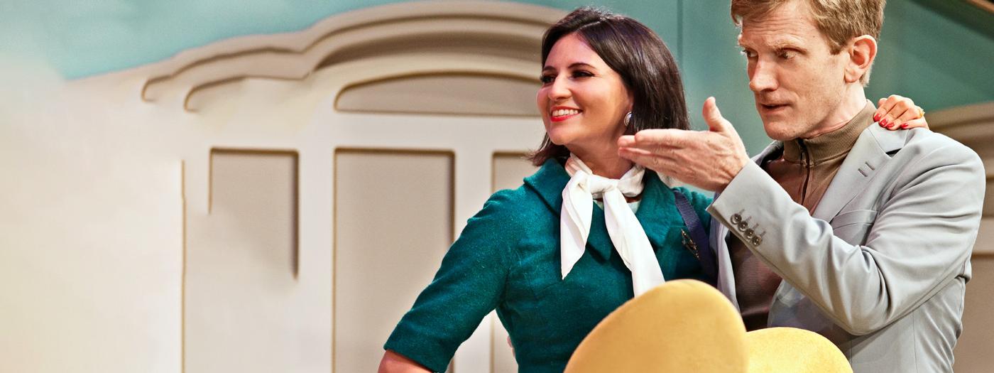 An air hostess in a dark green uniform and a white neck tie smiles with her arm around a man's shoulders, he is pointing and looking serious