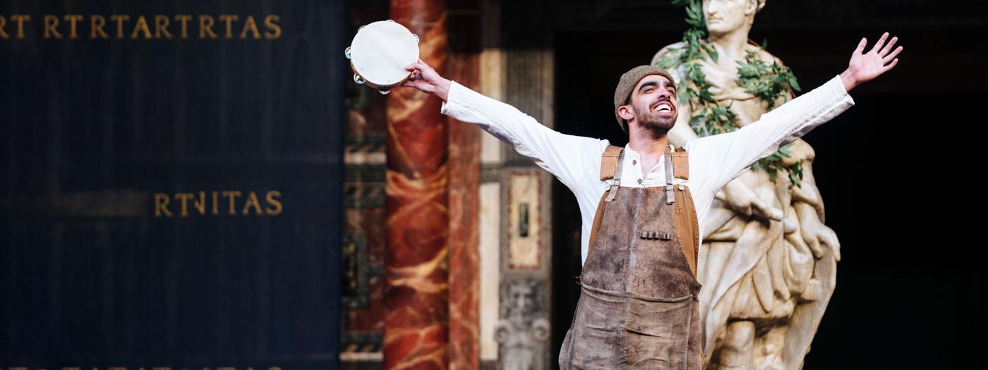 A man in a brown apron and beanie hat smiles with his arms outstretched, he holds a tambourine in one hand