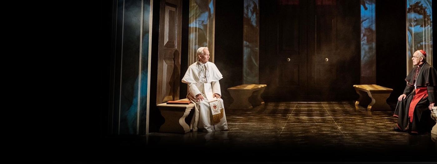 Two popes sit opposite each other on benches in a grand room