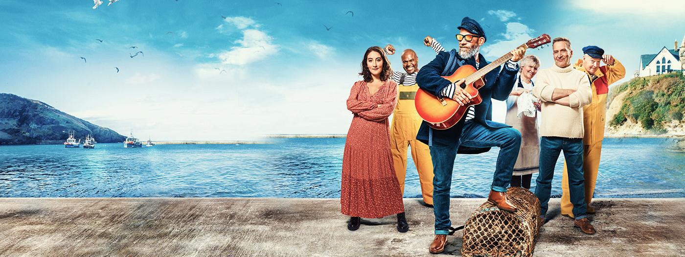 A fisherman with a guitar stands in front of the sea with a group of people