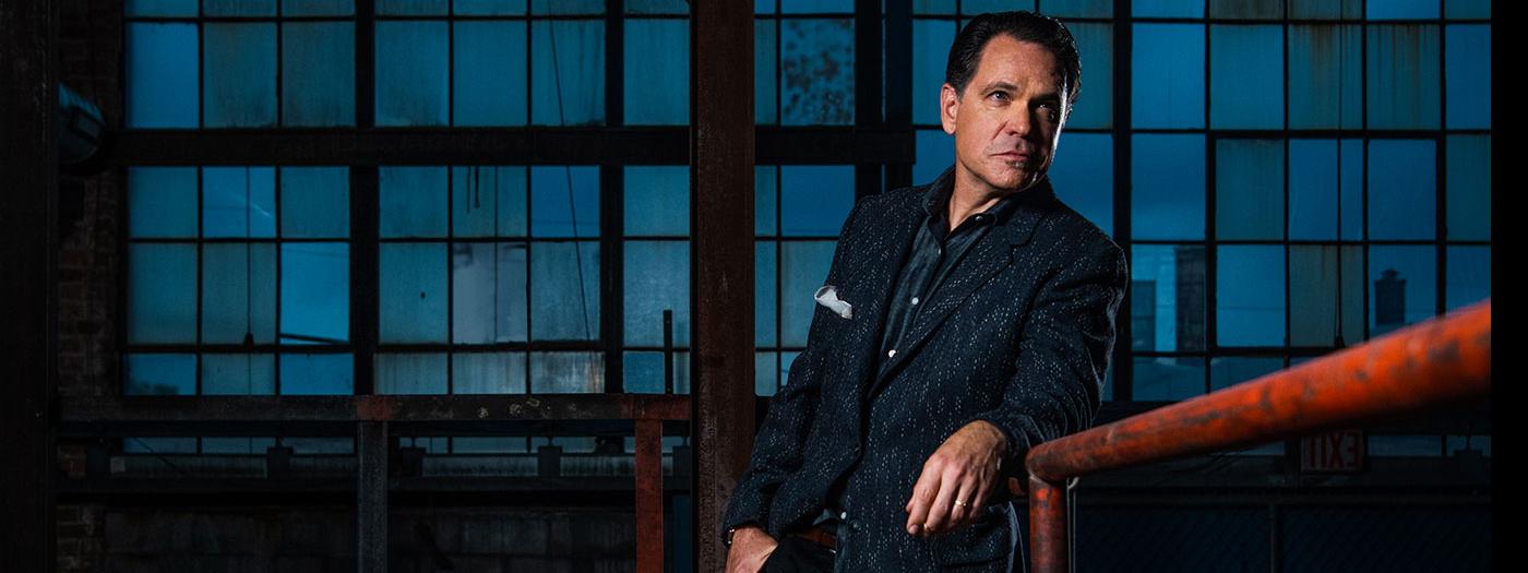 Kurt Elling stands by a metal rail, a blue window background is behind