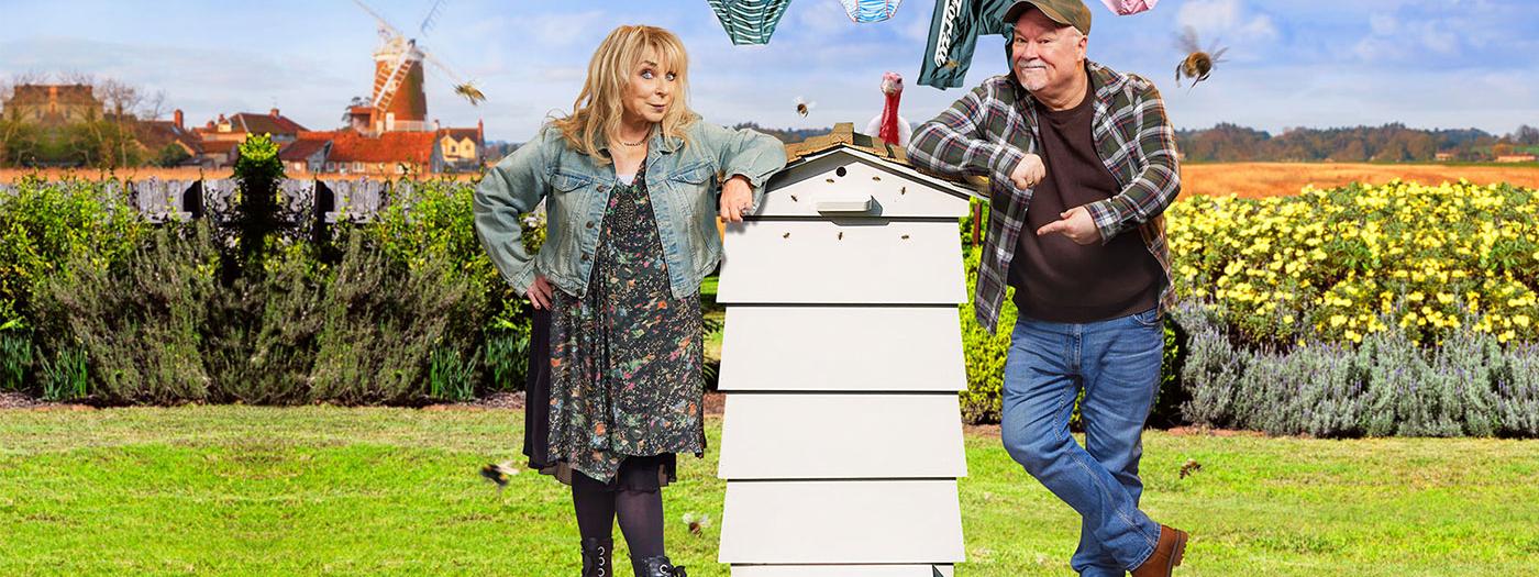 A woman and a man lean against a beehive, in the background there are fields and a washing line