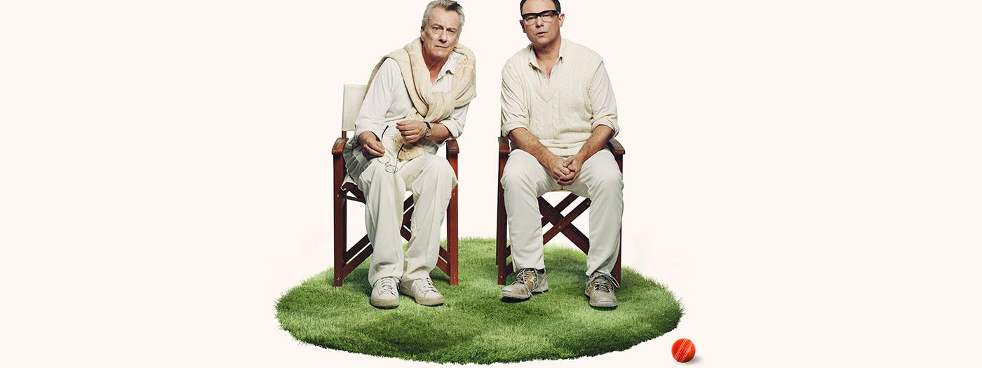 Two men wearing cricket gear sit in chairs on a small circular green