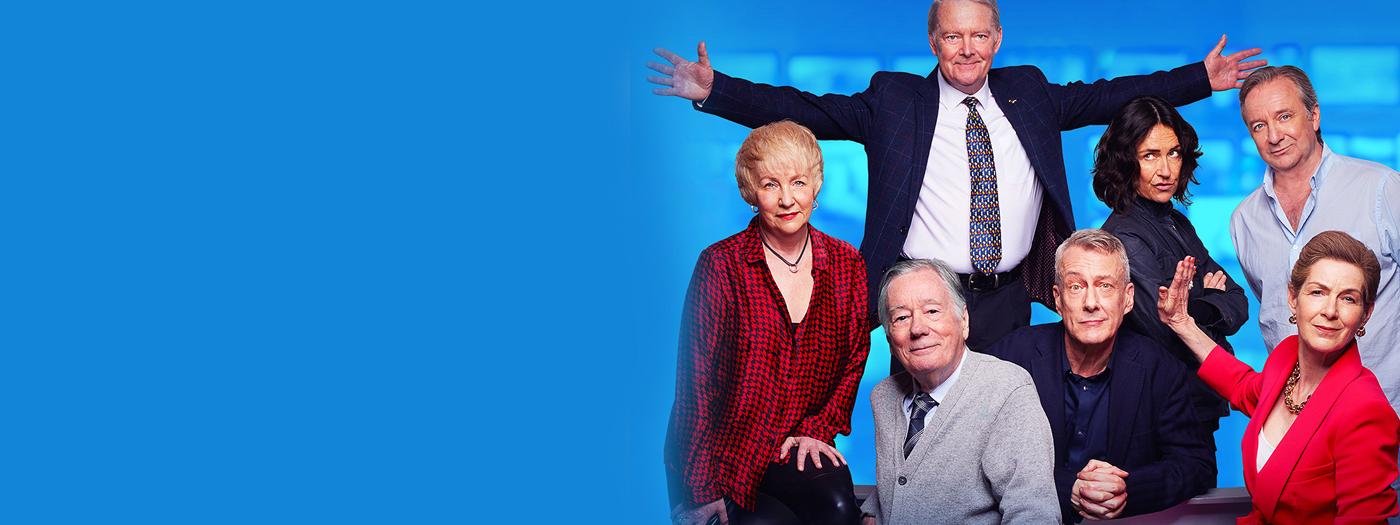 A group of people stand together around a newsdesk, one man has his arms outstretched and a woman holds her palm up against a man's face