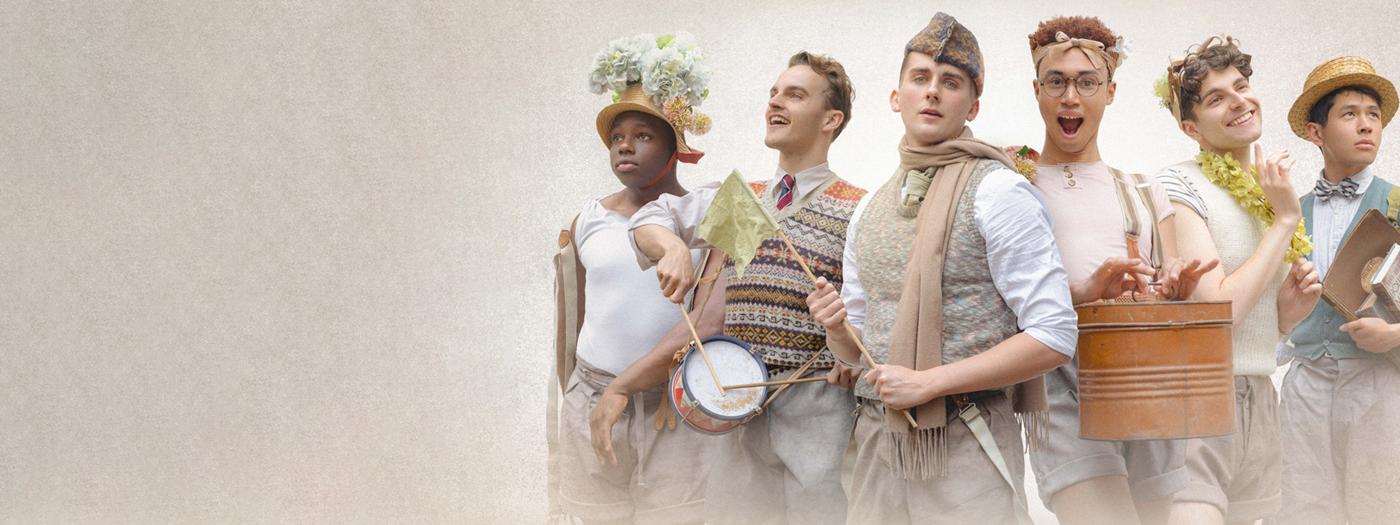 Six men stand together dressed in beige shorts and shirts, they have scarves, drums and flowered hats
