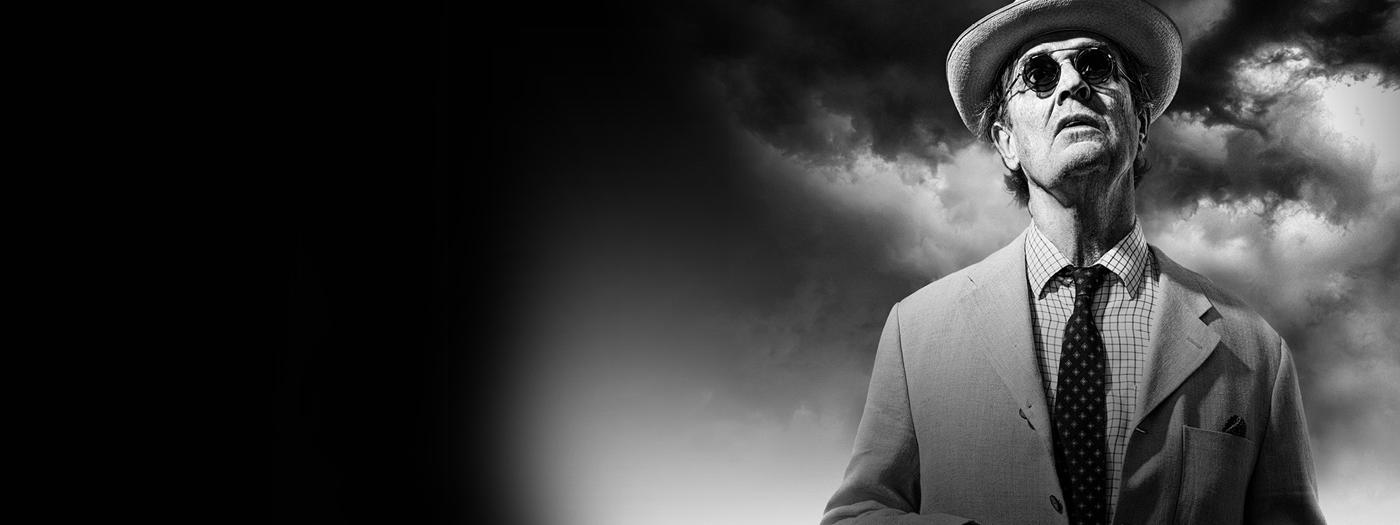 A man in a suit, hat and sunglasses stands against a cloudy sky