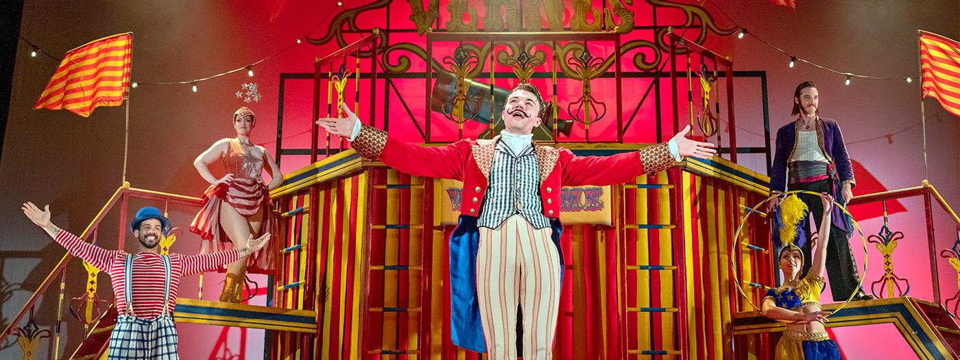 A circus scene, a man stands arms outstretched in a red ringleader outfit