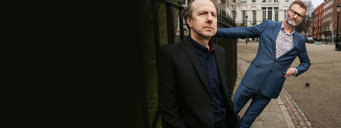 Two men in suits stand by some railings on a cobbled street