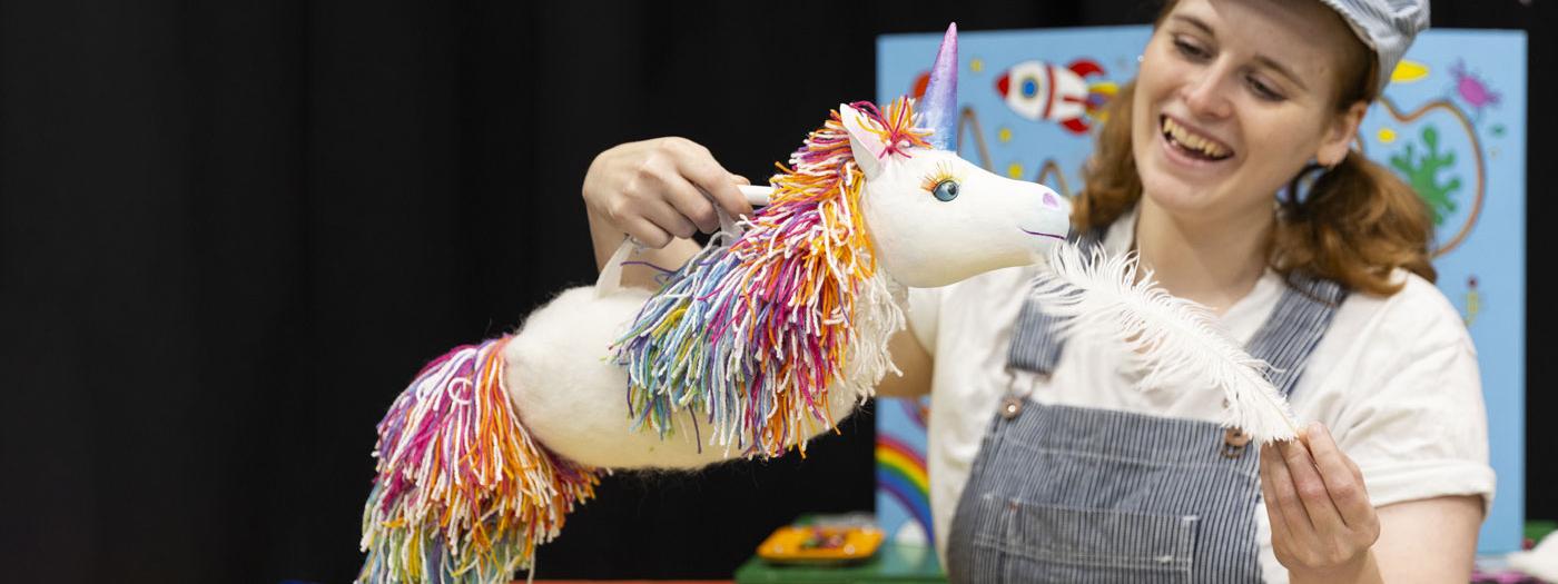 A woman wearing dungarees and a hat holds a unicorn and a big feather