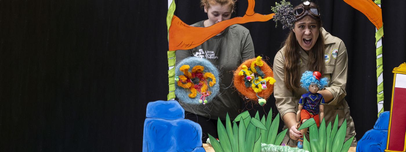 Two puppeteers in a jungle scene, one puppeteer is holding a puppet with blue hair